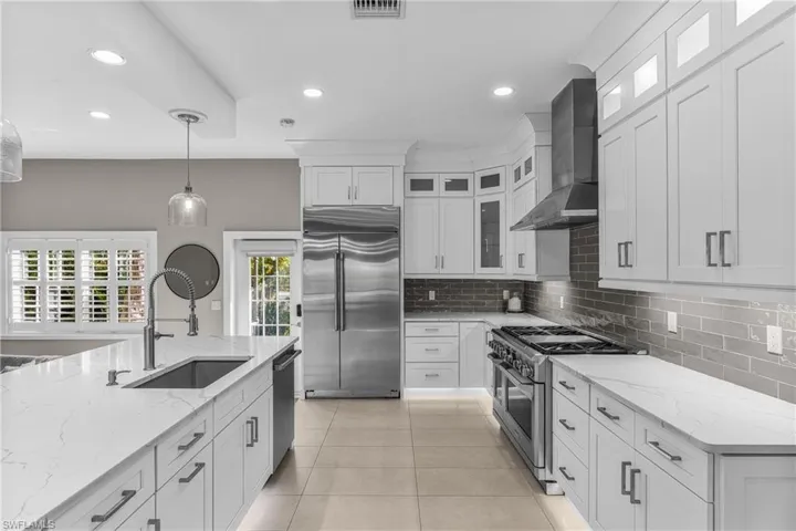 Kitchen featuring high end appliances, wall chimney range hood, light stone countertops, backsplash, and white cabinetry