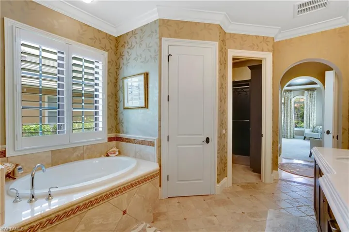 Primary Bathroom with wallpapered walls, ornamental molding, plenty of natural light, a soaking tub, and a large walk-in closet with custom shelving