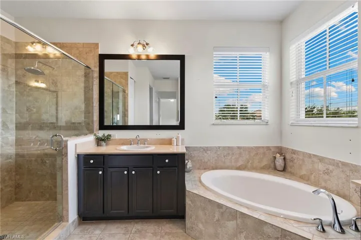 Spacious primary bathroom featuring an oval soaking tub, a separate glass-enclosed shower with a rainfall showerhead, a single vanity with a stone-finish countertop, and two large windows with horizontal blinds