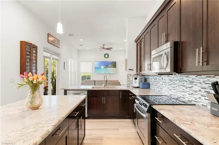 Open-concept kitchen featuring dark wood cabinetry, light-toned countertops, stainless steel appliances, a mosaic tile backsplash, and light wood-finish flooring