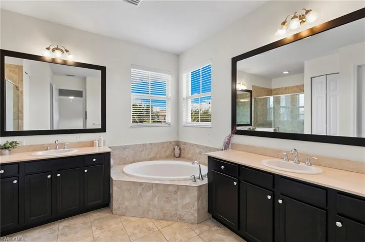 Primary Bathroom featuring a built-in soaking tub with tiled surround, dual vanity sinks set in light-toned countertops, dark wood-finish cabinetry with silver-tone hardware, and large framed mirrors with vanity lighting