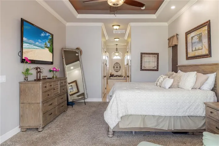 Bedroom featuring connected bathroom, light colored carpet, a raised ceiling, a ceiling fan, and ornamental molding