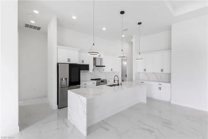 Kitchen featuring stainless steel appliances, light stone countertops, a kitchen island with sink, white cabinetry, and hanging light fixtures