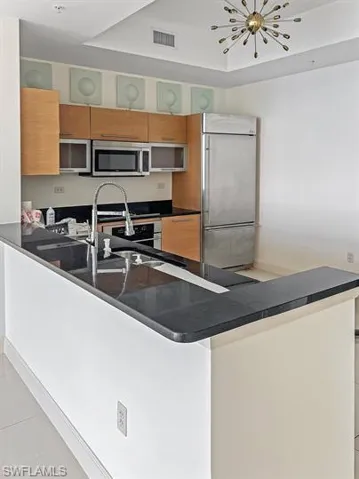 Kitchen with appliances with stainless steel finishes, a peninsula, light tile patterned floors, brown cabinetry, and a chandelier