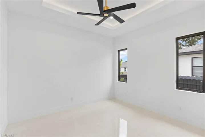 Tray ceiling with recessed lighting, ceiling fan with integrated light, two windows with dark frames, and light-toned tile flooring
