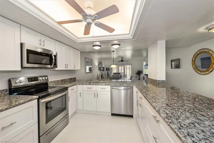 Kitchen featuring stainless steel appliances, a ceiling fan, a raised ceiling, white cabinets, and dark stone counters