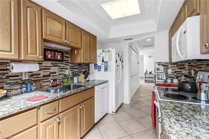 Kitchen with light stone counters, sink, white appliances, a tray ceiling, and crown molding