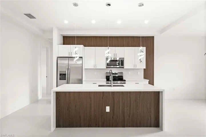 Kitchen featuring stainless steel appliances, light stone countertops, hanging light fixtures, white cabinetry, and a center island with sink