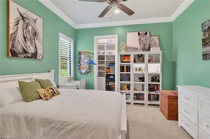 Bedroom featuring crown molding, neutral carpeting, a ceiling fan, a window with plantation shutters, and built-in shelving