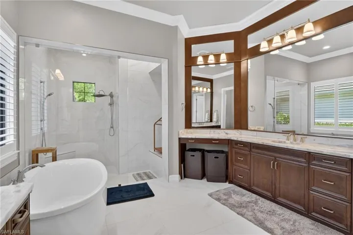 Spacious bathroom featuring a freestanding soaking tub, a large glass-enclosed shower with multiple showerheads, a dual-sink vanity with extensive counter space, custom wooden cabinetry, and substantial mirror fixtures with integrated lighting