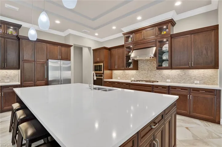 Expansive kitchen island with white countertop, rich wood-finish cabinetry, stainless steel appliances, mosaic tile backsplash, and decorative ceiling details