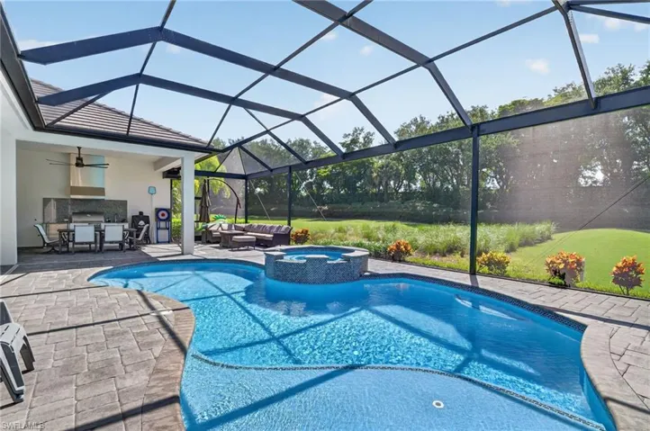Screened lanai featuring a freeform pool, integrated spa with mosaic tile, paver decking, and a built-in outdoor kitchen with a stainless steel range hood