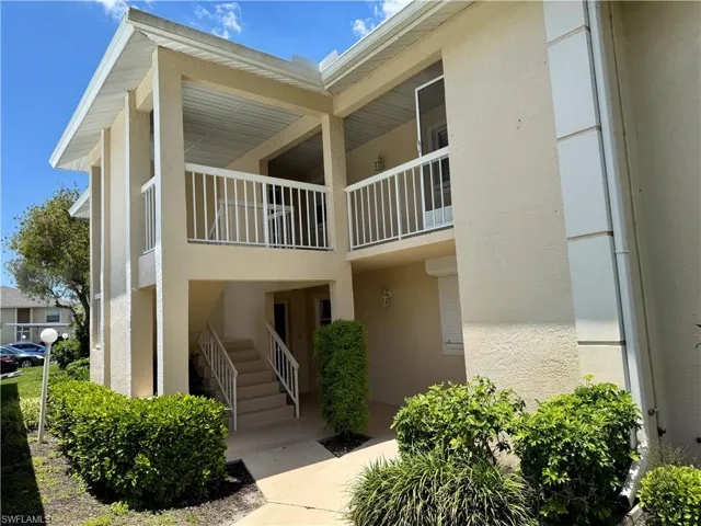 Two-story condominium building featuring stucco siding, multiple balconies with white railings, exterior sconce lighting, and mature landscaping