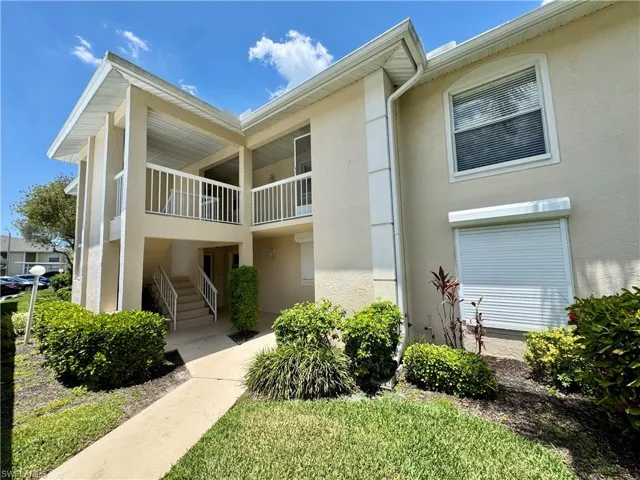 Multi-story condominium building featuring a neutral stucco exterior, covered balconies with white railings, and a ground-floor entrance with concrete steps