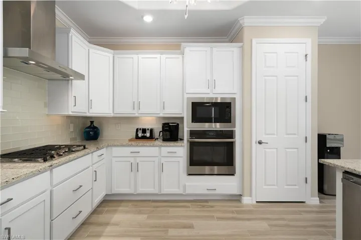 Kitchen with tasteful backsplash, stainless steel appliances, wall chimney exhaust hood, white cabinetry, and ornamental molding