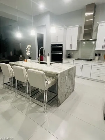 Kitchen featuring wall chimney exhaust hood, light stone counters, a kitchen bar, stainless steel double oven, and white cabinets