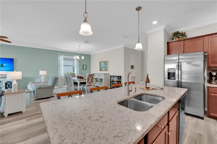Kitchen with stainless steel fridge, ornamental molding, light stone counters, brown cabinetry, and decorative light fixtures