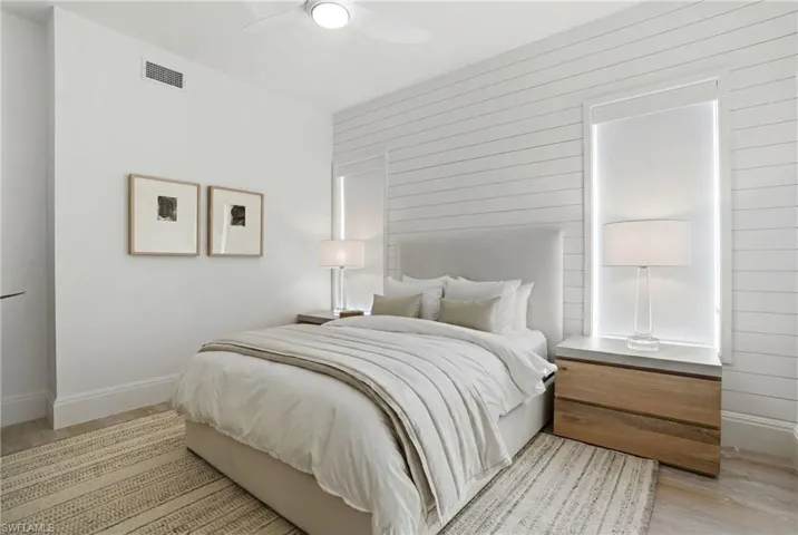 Upholstered headboard, wood-finish flooring, accent wall with horizontal paneling, ceiling fan with integrated lighting, and a window with roller shade