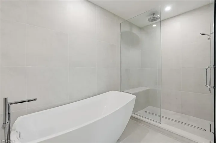 Freestanding soaking tub with chrome fixtures, large format tile walls, glass-enclosed shower, rain shower head, and built-in shower bench
