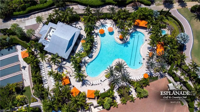 Curvilinear swimming pool with integrated lap lanes, expansive pool deck with multiple lounge chairs, surrounding mature palm trees, adjacent building with a metal roof, and nearby tennis courts