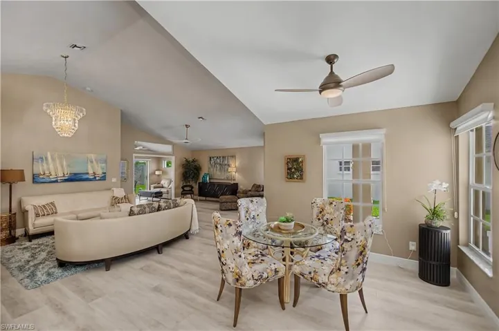 Spacious open-concept living area featuring vaulted ceilings, light wood-finish flooring, a contemporary crystal chandelier, and a ceiling fan with integrated lighting