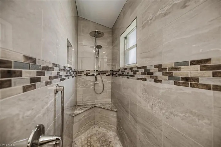 Spacious shower featuring large format light-toned wall tiles, a multi-hued horizontal accent tile band, a built-in corner bench, a recessed shower niche, and a contemporary rainfall showerhead with a handheld sprayer