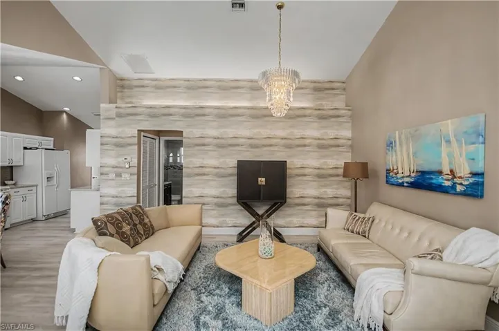 Vaulted ceiling living area featuring a decorative chandelier, accent wall, wood-finish flooring, and a partial view of the kitchen with white cabinetry