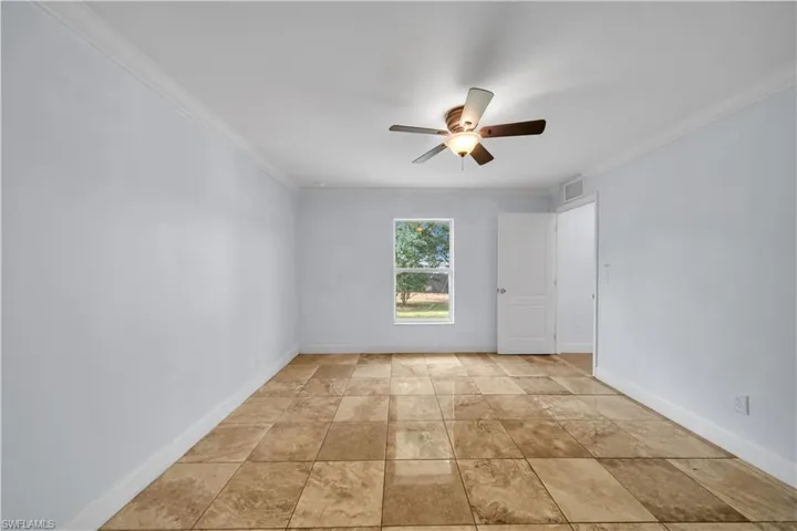 Room featuring large format tile flooring, crown molding, a ceiling fan with integrated lighting, and a single window with exterior landscaping visible