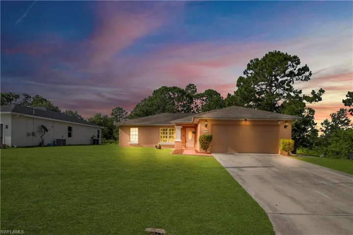 Ranch-style residence featuring a tan stucco exterior, a two-car garage with overhead lighting, a paved driveway, a covered entry with a white paneled door, and a manicured front lawn