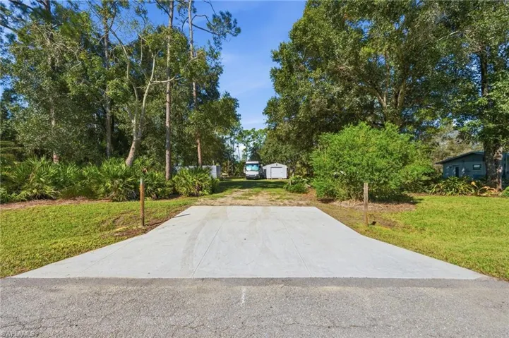 Concrete driveway apron opening to an expansive lot featuring mature trees, native landscaping, and a detached storage structure