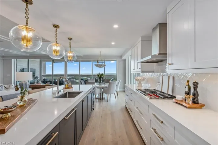 Kitchen with white cabinets, hanging light fixtures, light wood-style floors, light stone countertops, and wall chimney exhaust hood