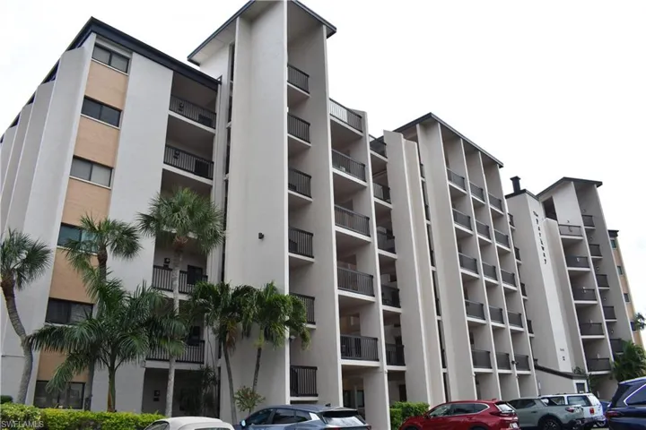 Multi-story building featuring numerous private balconies with dark railings, a white and tan exterior facade, and mature palm trees at the ground level