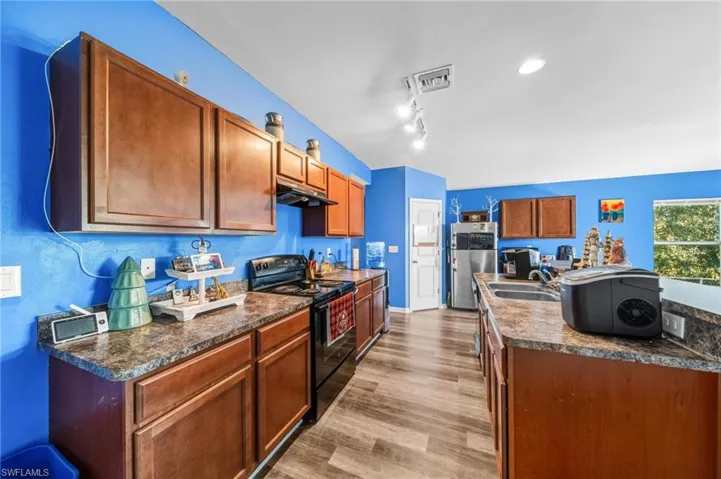 Kitchen with dark countertops, black electric range oven, a kitchen island with sink, stainless steel refrigerator, and brown cabinets