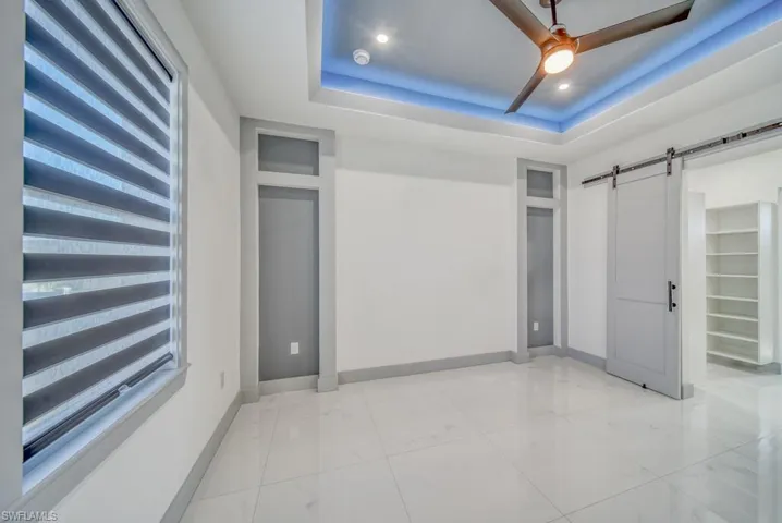 Unfurnished bedroom featuring a barn door, baseboards, a tray ceiling, light marble finish flooring, and ceiling fan