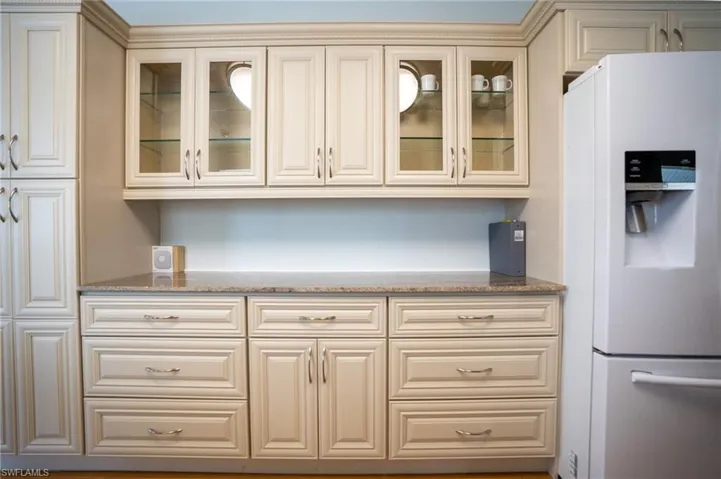 Kitchen featuring white refrigerator with ice dispenser, cream cabinets, light stone countertops, and glass insert cabinets