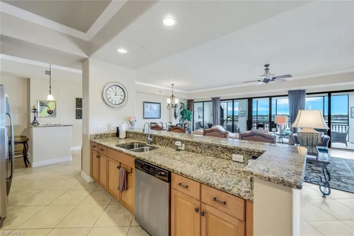 Kitchen with breakfast bar, light tile patterned floors, ceiling fan with notable chandelier, light stone counters, appliances with stainless steel finishes, facing Grand room and screened lanai.
