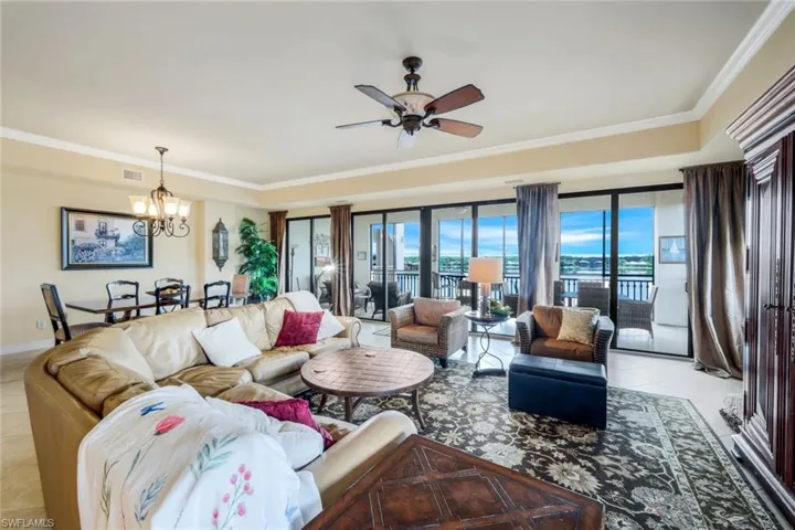 Diagonally Tiled grand room with ceiling fan with notable chandelier and ornamental crown molding! Facing wall of impact glass sliders over looking large screened lanai overlooking beac, lake and beach club.