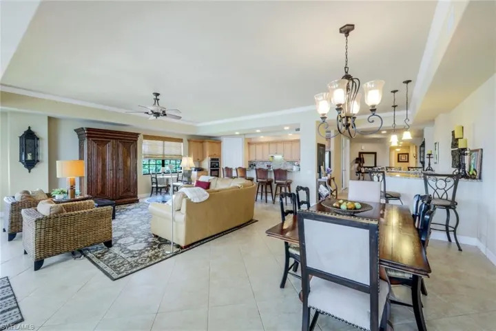 Grand room facing kitchen and wet bar featuring ceiling fan with notable chandelier, ornamental molding, light tile patterned floors, and a tray ceiling