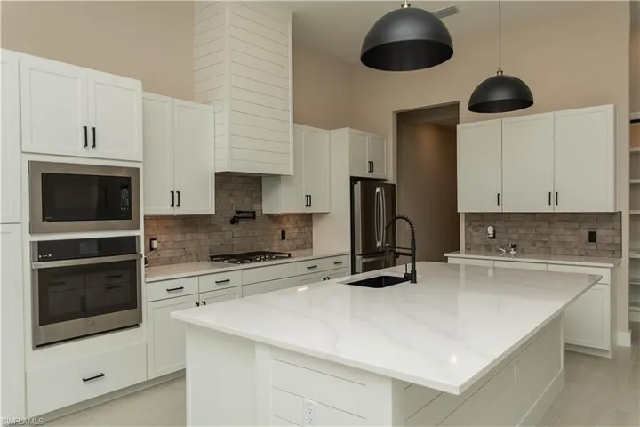 Kitchen with white cabinets, an island with upgraded matte black sink, quartz countertops, pot filler, tile backsplash, upgraded pantry, decorative light fixtures, and stainless steel appliances,  views of pool/spa and golf course lake.