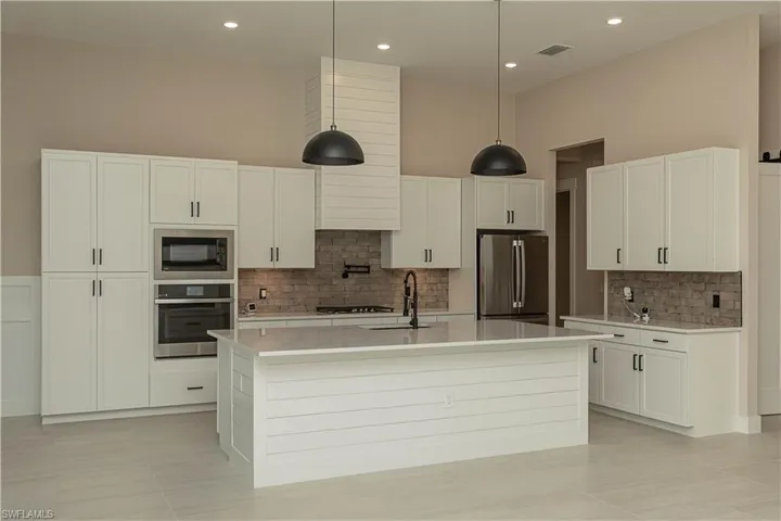 Kitchen with white cabinets, an island with upgraded matte black sink, quartz countertops, pot filler, tile backsplash, upgraded pantry, decorative light fixtures, and stainless steel appliances,  views of pool/spa and golf course lake.