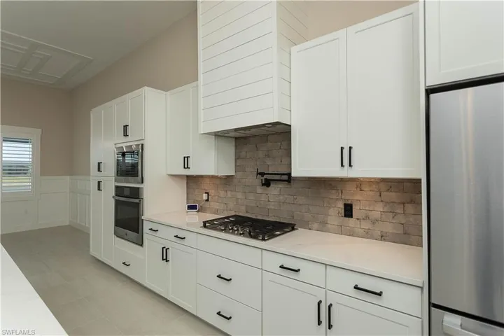 Kitchen with white cabinets, an island with upgraded matte black sink, quartz countertops, pot filler, tile backsplash, upgraded pantry, decorative light fixtures, and stainless steel appliances,  views of pool/spa and golf course lake.