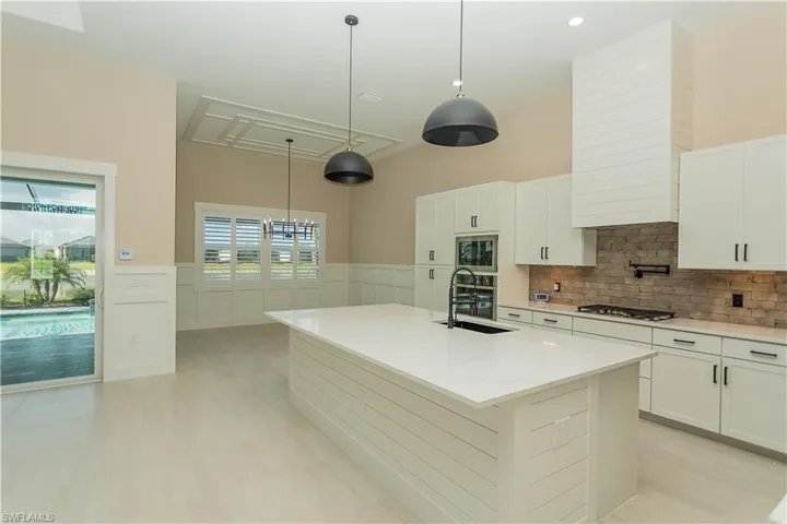 Kitchen with white cabinets, an island with upgraded matte black sink, quartz countertops, pot filler, tile backsplash, upgraded pantry, decorative light fixtures, and stainless steel appliances,  views of pool/spa and golf course lake.