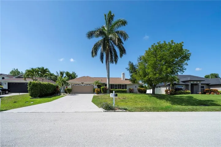 Single-story residence featuring a two-car garage with a beige door, an asphalt driveway, a front lawn with mature trees and shrubbery, and a prominent palm tree