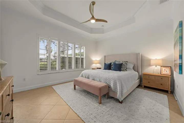 Primary bedroom with a tray ceiling with crown molding and modern ceiling fan, large window with plantation shutters, and tile flooring