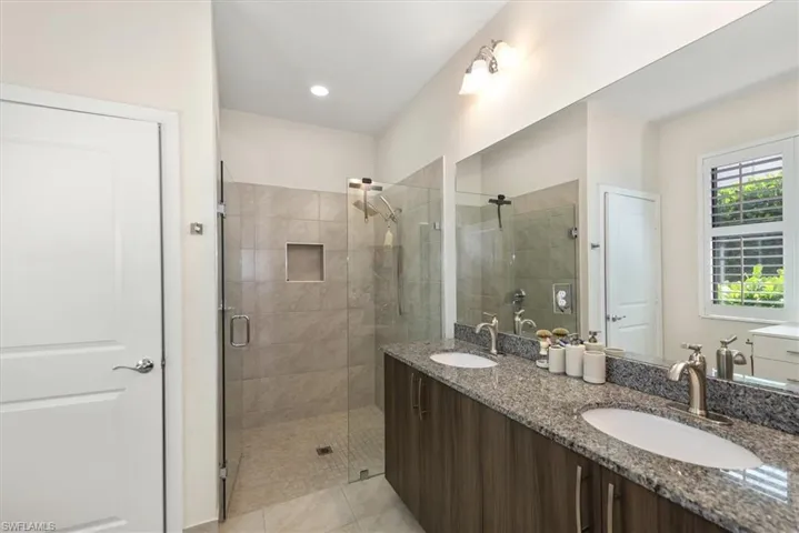 Bathroom featuring a dual vanity with granite countertops and undermount sinks, a large frameless mirror, a glass-enclosed shower with tiled walls and an integrated niche, and a window with plantation shutters