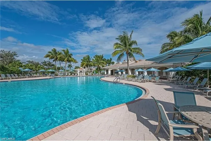 Resort-style pool area featuring a large, sparkling blue pool, extensive paver decking, numerous lounge chairs, and shade umbrellas