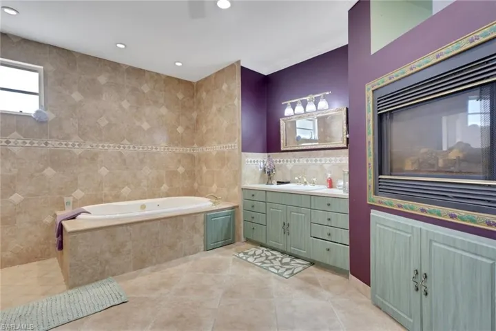Bathroom featuring a built-in jetted tub, extensive tilework, a double vanity with light green cabinetry, and an integrated fireplace