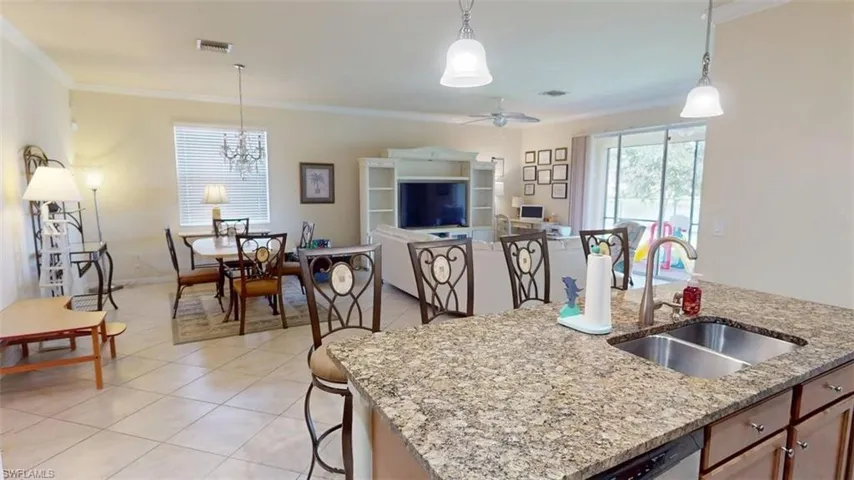 Kitchen with pendant lighting, light tile patterned floors, crown molding, light stone counters, and a kitchen island with sink
