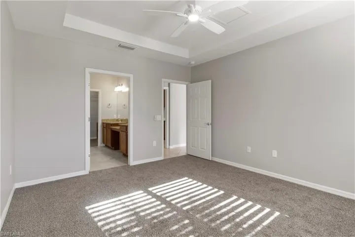 Unfurnished bedroom with a tray ceiling, light colored carpet, ensuite bath, and ceiling fan