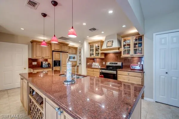 Kitchen featuring a spacious island, built in appliances, custom range hood, backsplash, and decorative light fixtures